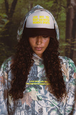 Person wearing a camouflage hoodie and cap with text in a forest setting