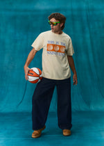 A man wearing a cream graphic t-shirt with blue and orange text, dark navy wide-leg pants, and brown boots, holding a basketball against a textured blue backdrop.