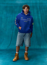 A man stands against a teal studio backdrop wearing a royal blue hoodie with yellow and white graphics, light-wash denim shorts, white socks, and tan lace-up boots. The hoodie has 'ORIGINALS' printed across the chest and '1996' on the sleeve.