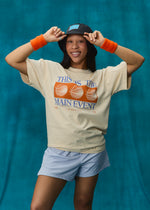 A woman posing in a beige graphic t-shirt with orange and blue text and basketball graphics, paired with light blue shorts, orange wristbands, and a black bucket hat against a teal textured backdrop.