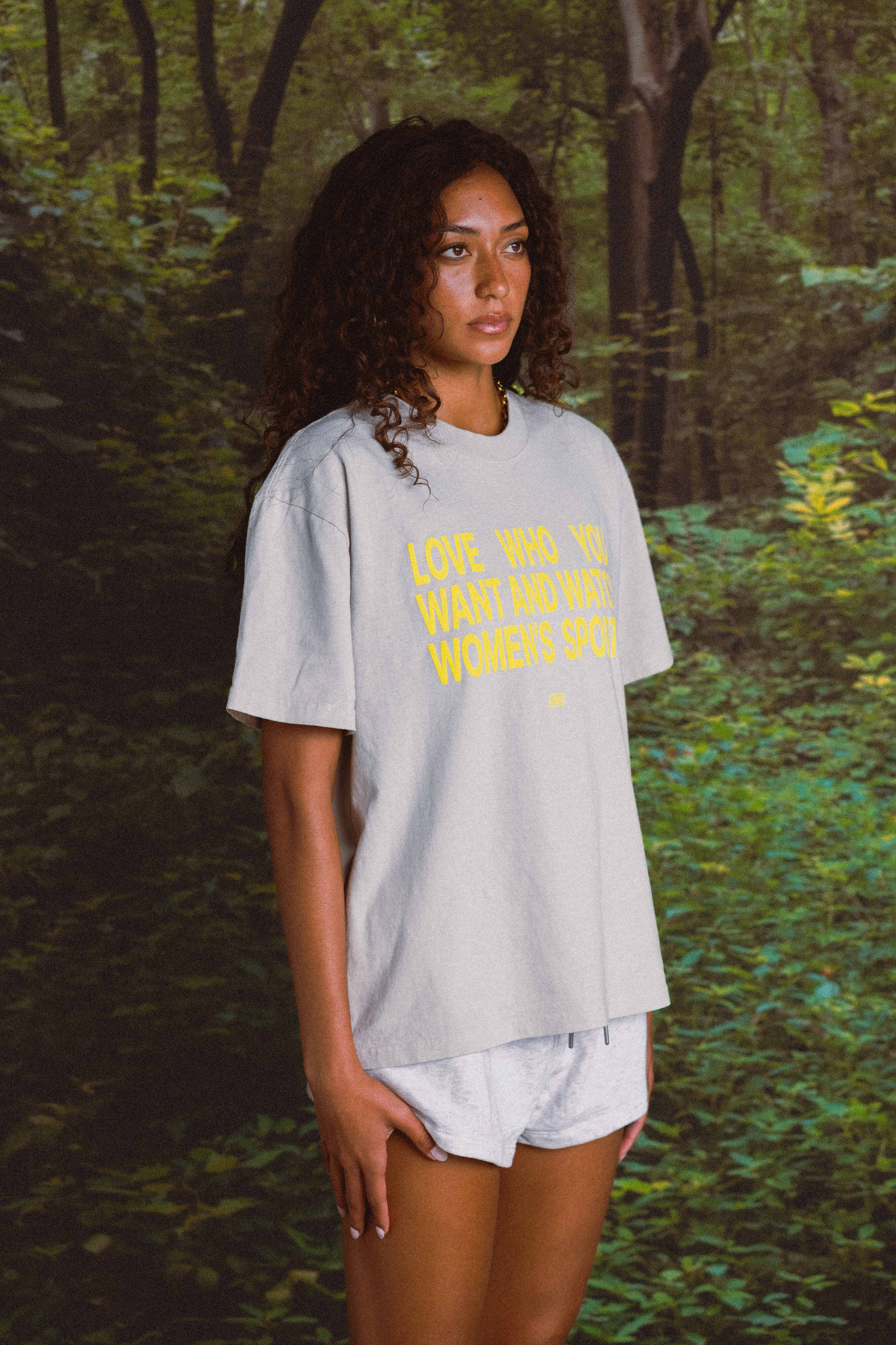 Woman wearing a light gray t-shirt with yellow text in a forest setting