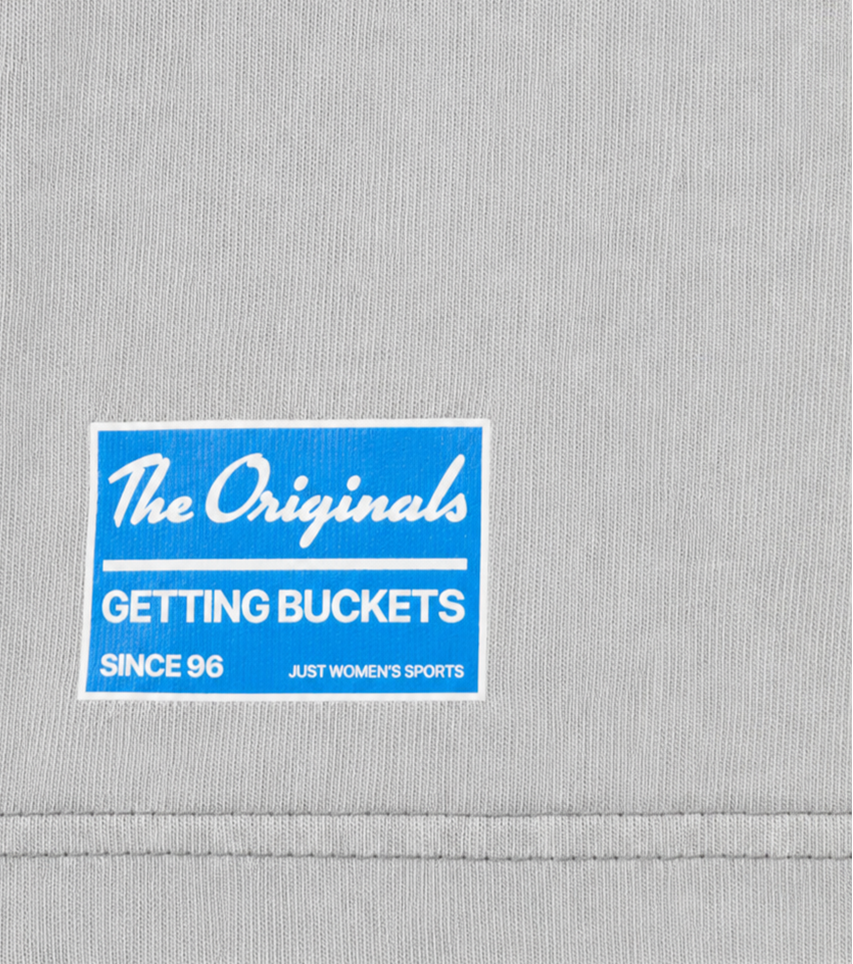 Close-up of a light gray ribbed fabric sleeve with a blue and white rectangular printed patch reading 'The Originals GETTING BUCKETS SINCE 96 JUST WOMEN'S SPORTS'.