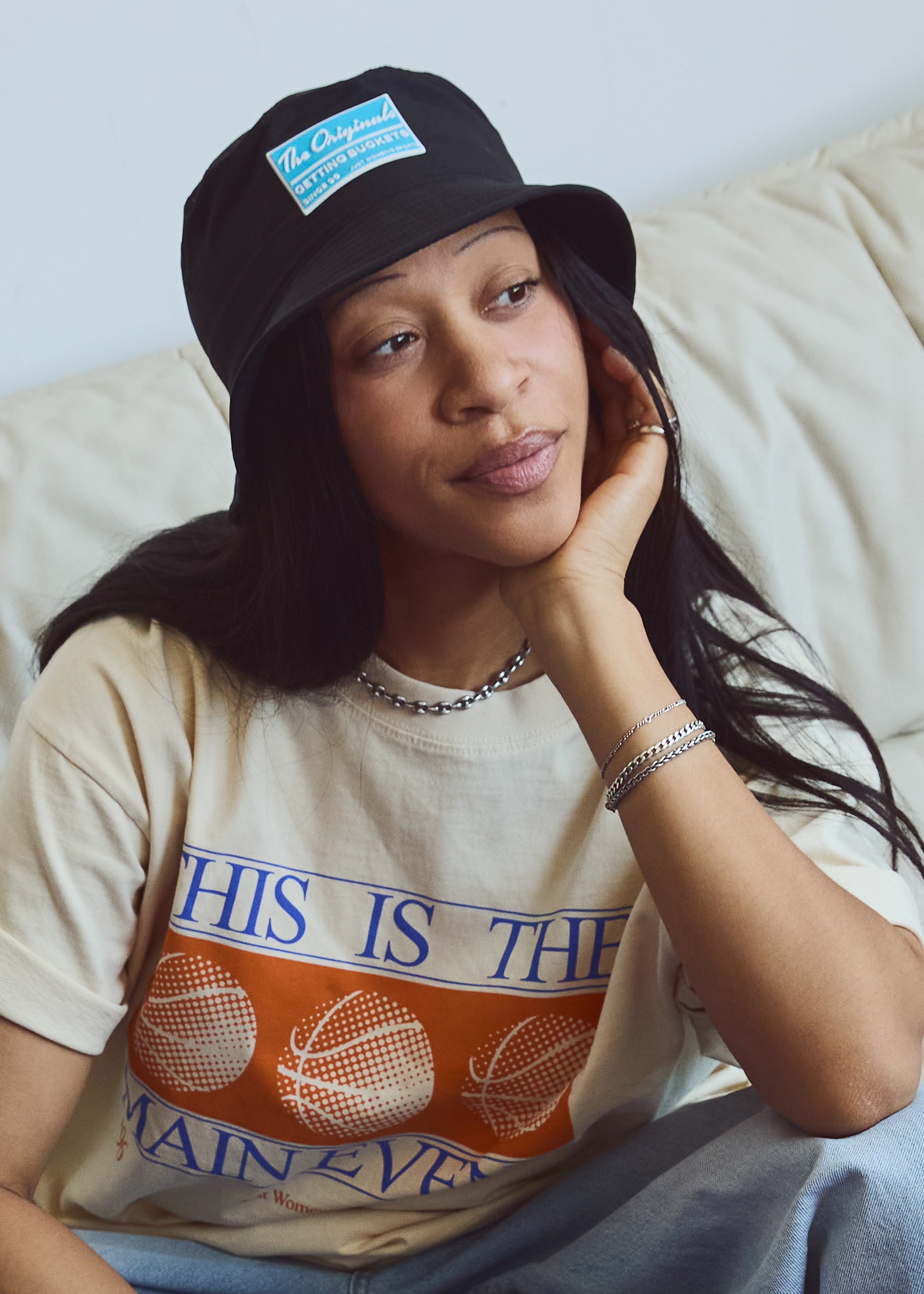 A woman wearing a black bucket hat with a blue and white 'The Originals' patch and a cream graphic t-shirt with orange and blue text, sitting on a beige couch.